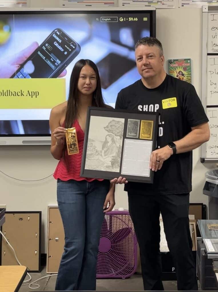 The 1st place winner and a Goldback representative stand in an art classroom holding a framed artwork display and a Goldback note. Behind them, a classroom screen shows the Goldback App page.
